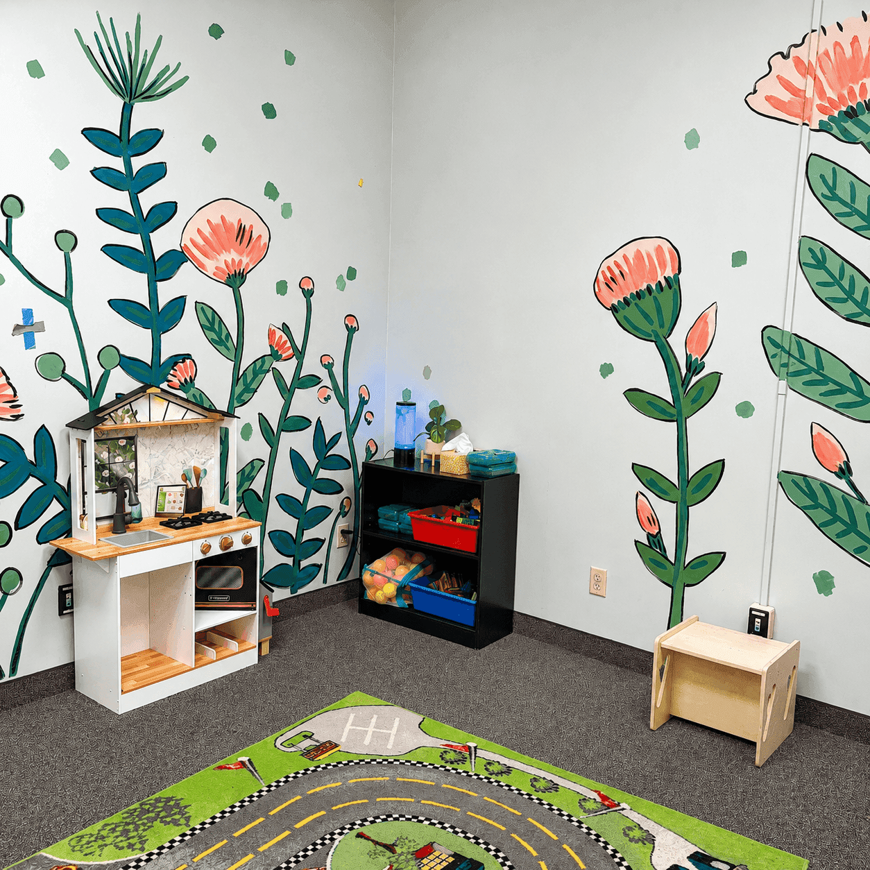 A play corner with a botanical wall mural, a wooden toy kitchen, toy bins, and a road-and-car play rug