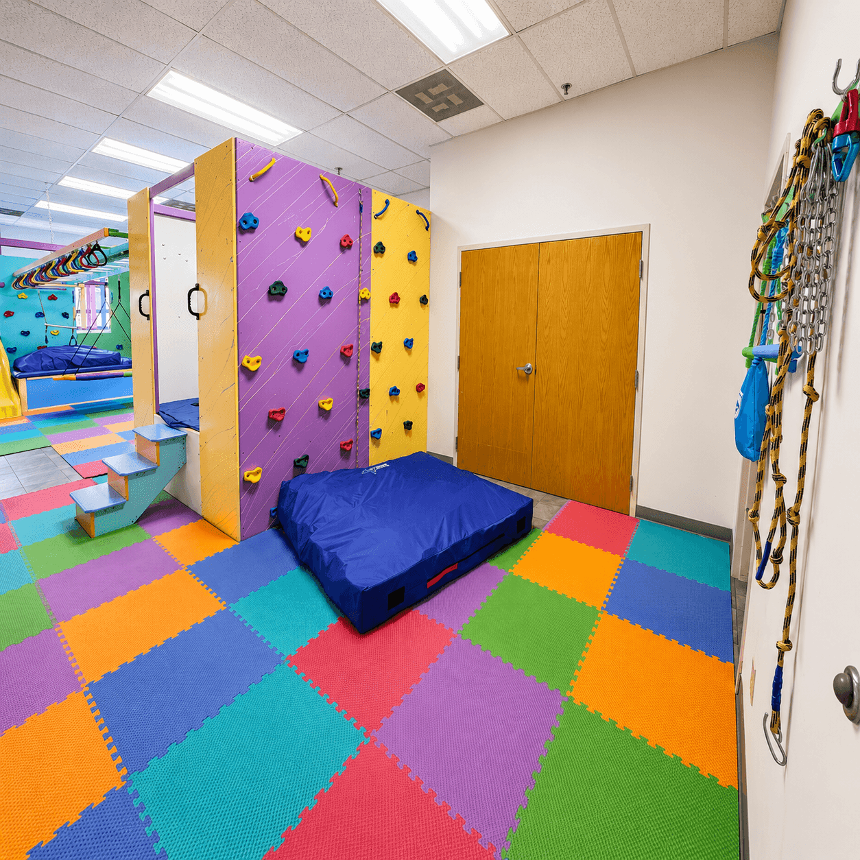 A sensory gym with a colorful climbing wall, a padded crash mat, and rope ladders over a soft foam-tile floor