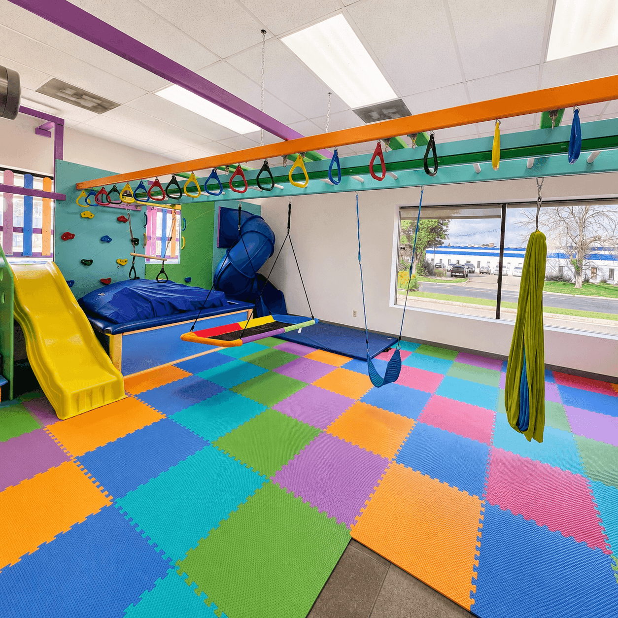 A bright indoor therapy gym filled with colorful sensory and motor equipment over a soft padded floor